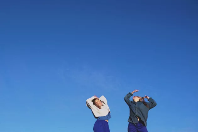 Foto: två dansande kvinnor med uppsträckta armar mot en blå himmel.
