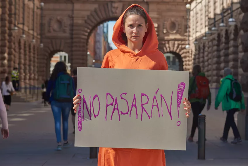 Kvinna i orange tröja håller upp en skylt framför sig med ett textbudskap. Foto