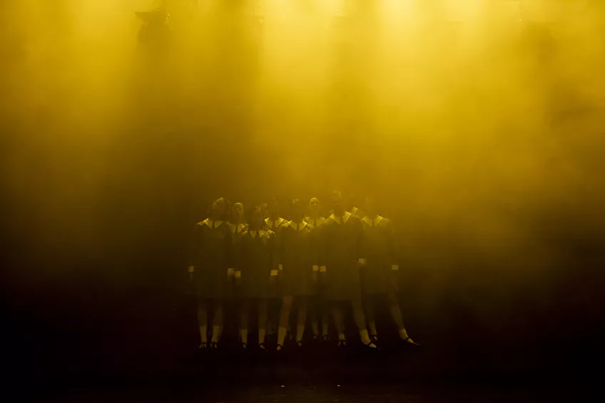 Foto på skådespelare i likadana skor, strumpor och klänningar i gult ljus och rök.