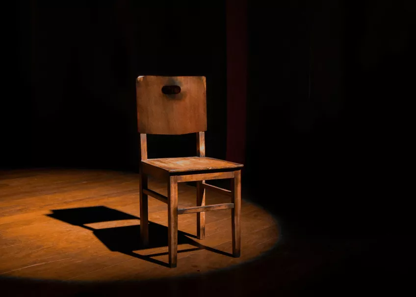 En stol av trä, upplyst av en spotlight på en mörk scen. Fotografi.